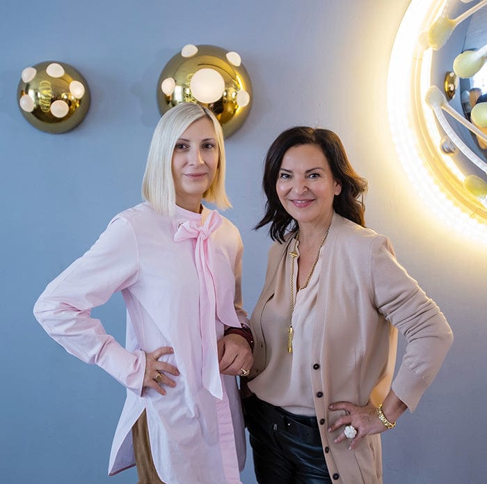 Veronique and Grimanesa posing in front of a white wall with gold accents.