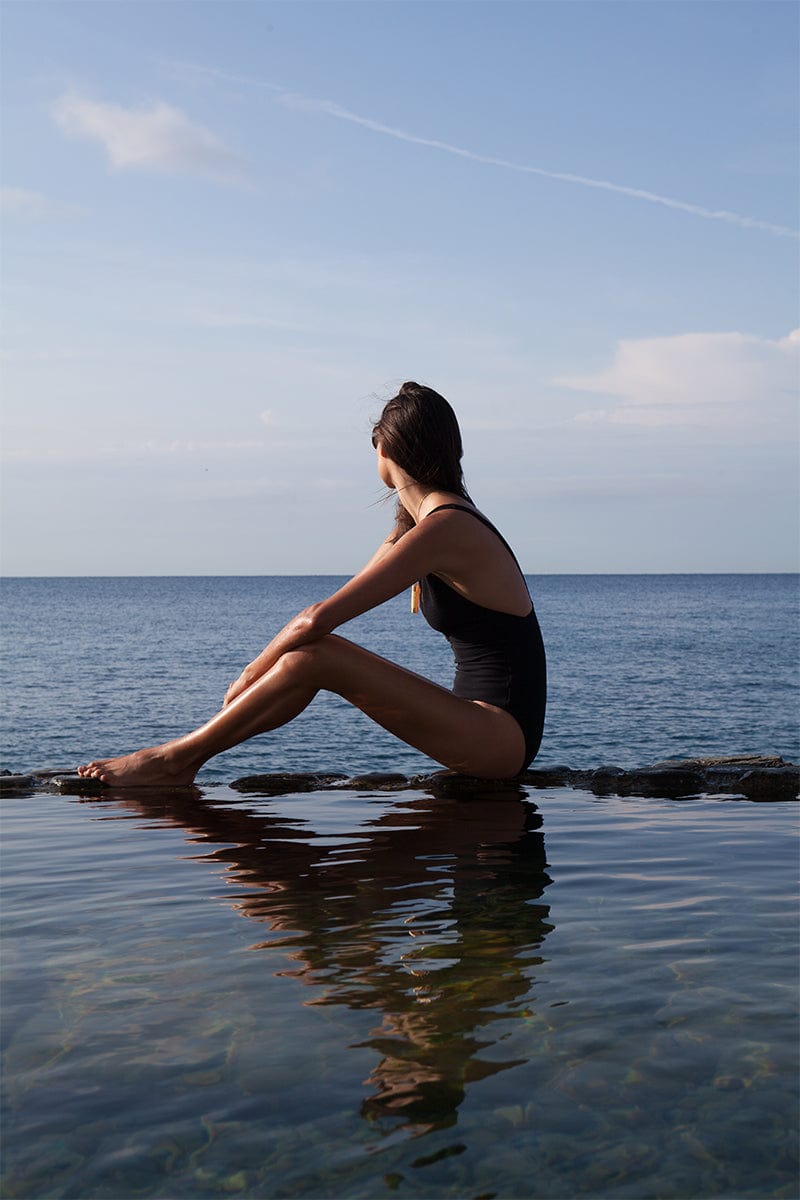 Woman sitting by the rocky beach wearing a black swimsuit and Veronique Gabai's Le Spritz pendant in Gold. 